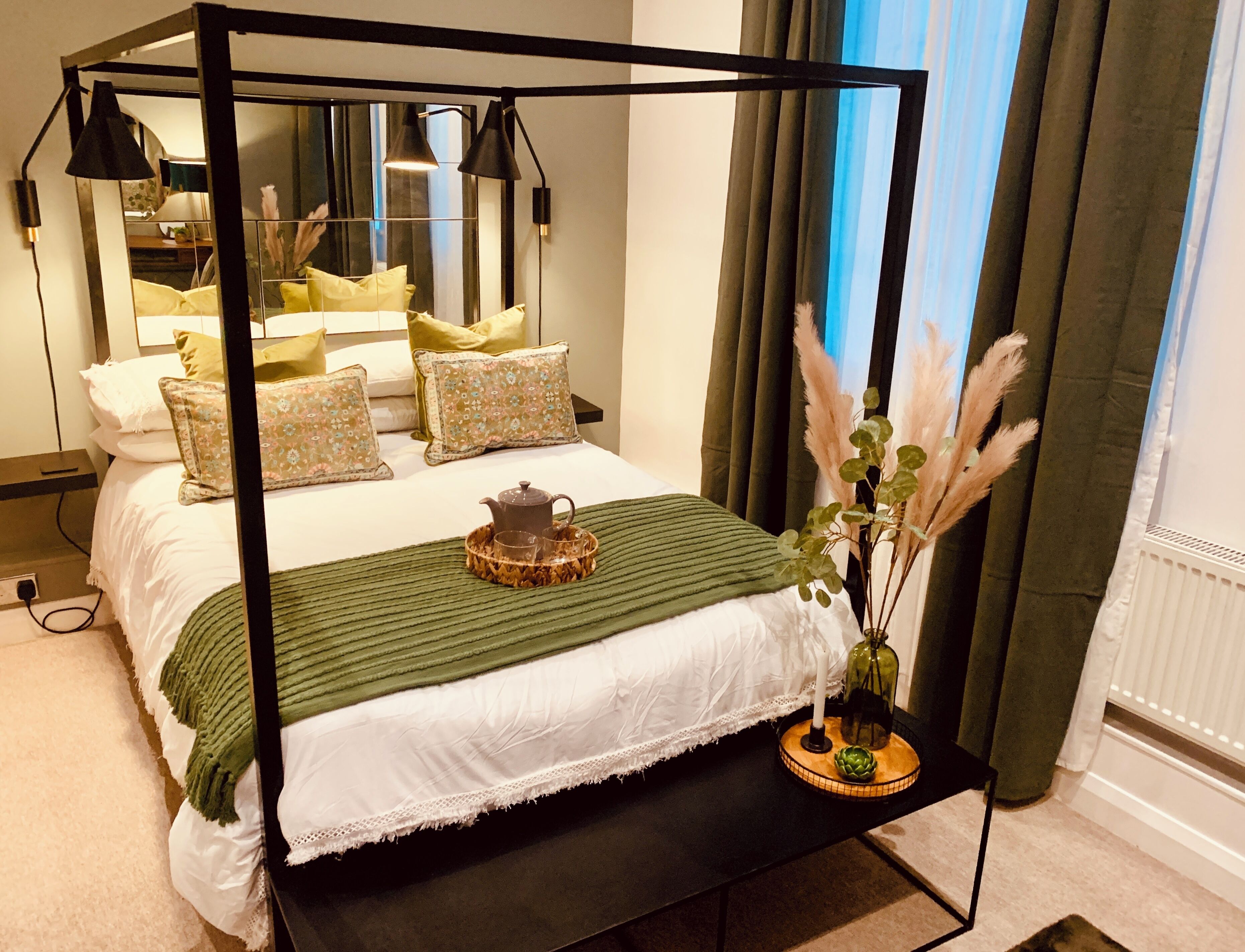 Un lit à baldaquin surmonté d’un plaid vert, une table de chevet avec une bougie et un vase rempli de plumes d’herbe des pampas, et un miroir qui reflète la pièce.