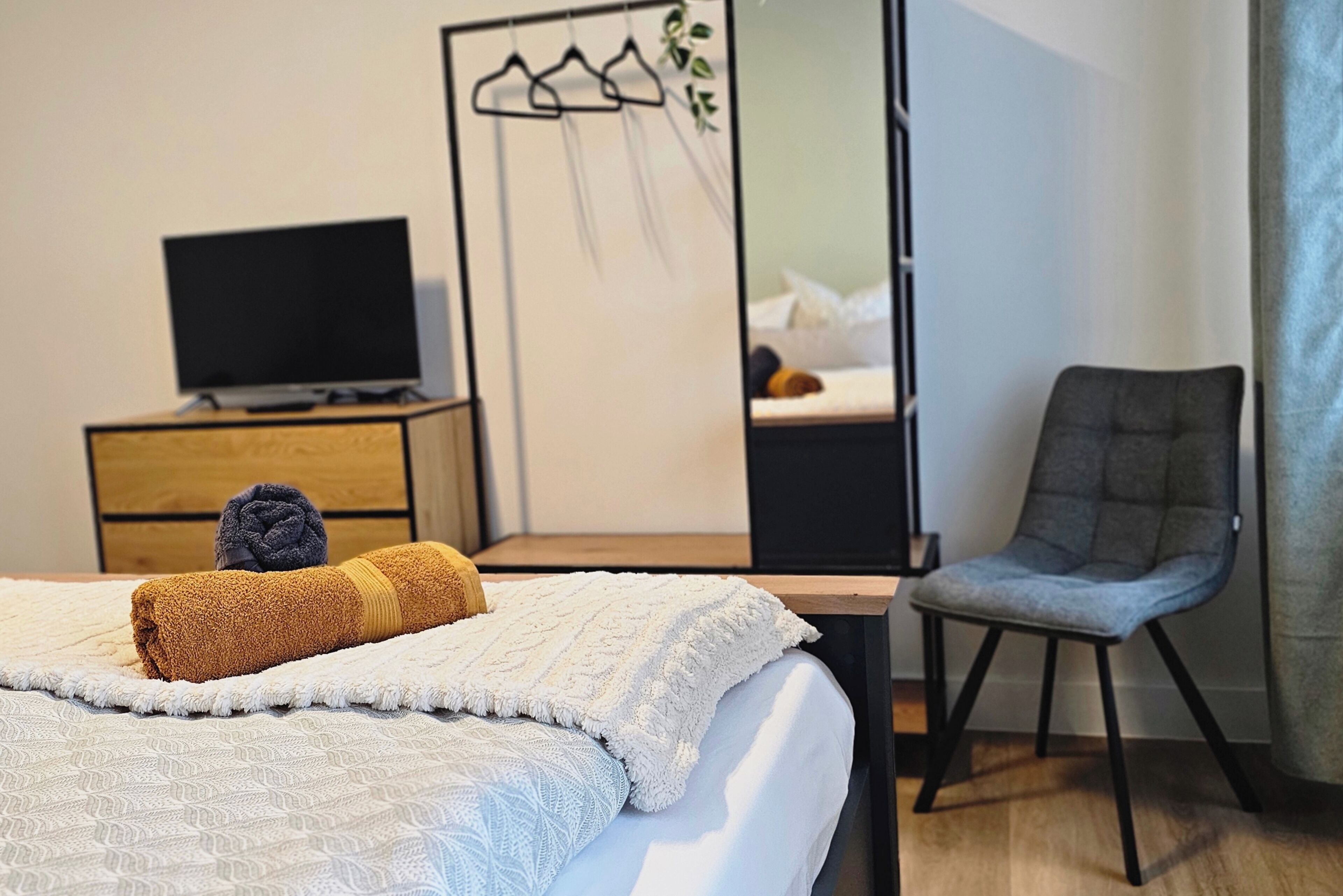 Ein ordentlich bezogenes Bett mit weißer und gemusterter Decke, ein grauer Sessel, ein Fernseher auf einer Holzkommode und ein Spiegel mit einer Pflanze.