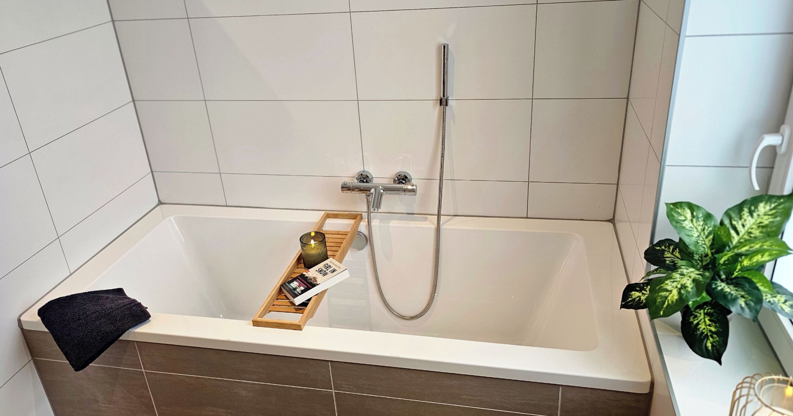 A modern bathroom with a bathtub, a wooden tray with books and a candle, and a potted plant.