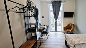 A bedroom with a bed, a desk with a chair, a hanging plant, and a TV on the wall.