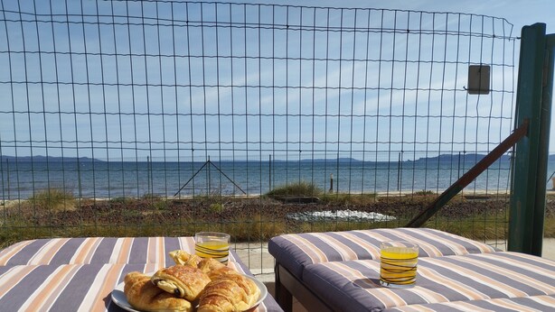 Una cama junto a la playa con ropa de cama a rayas, un plato de croissants y dos vasos de zumo de naranja, con vistas al mar.