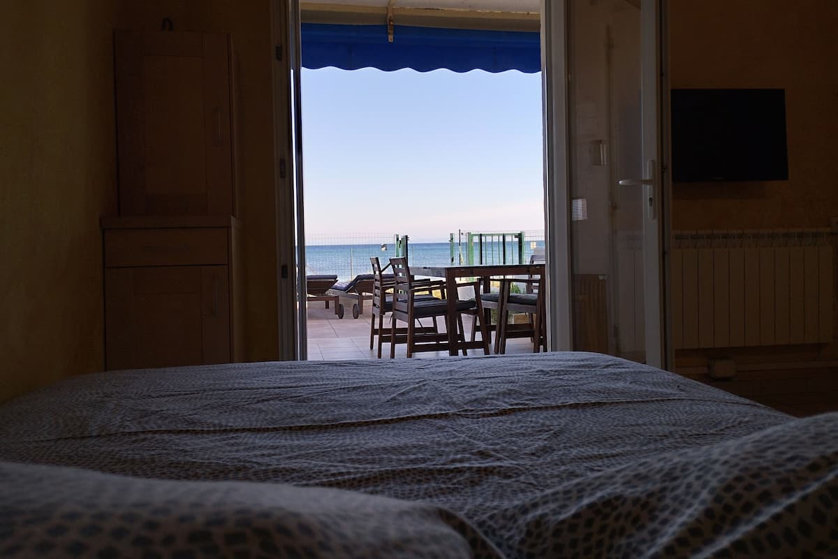 A room with a bed, a view of the beach, and a table with chairs outside.