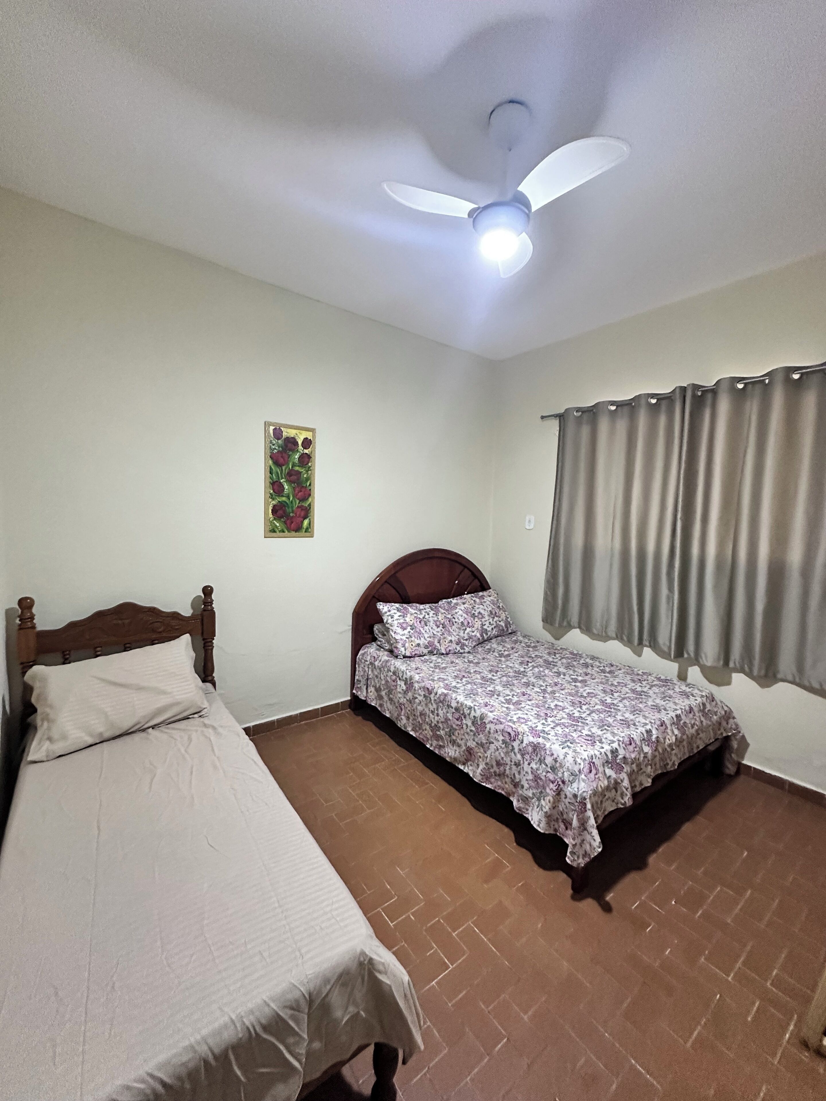 Une chambre avec deux lits, un ventilateur de plafond, une fenêtre avec des rideaux et un tableau au mur.