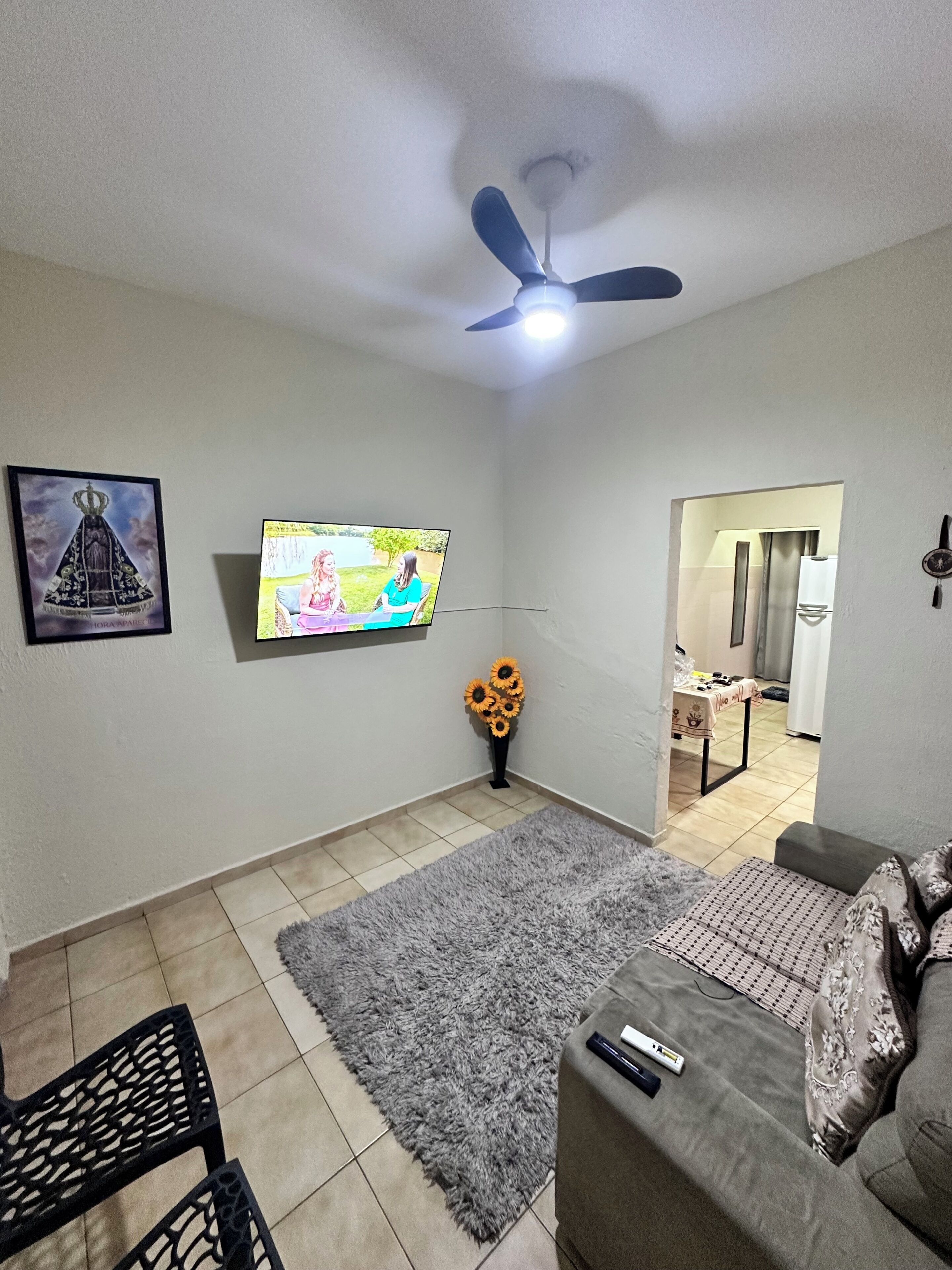 Una sala de estar con televisor, ventilador de techo, alfombra gris, sofá y una mesita con un jarrón de girasoles.