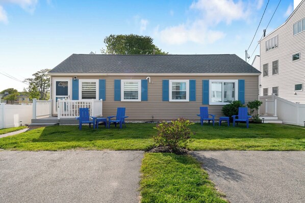 Une maison à deux étages avec un porche à l’avant, des volets bleus et une petite pelouse agrémentée de quatre chaises bleues.