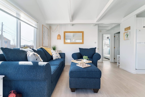 A modern living room with a blue sofa and ottoman, a wooden-framed mirror, and a view through a large window.