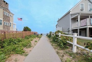 A coastal walkway with a wooden fence, a sandy path, and a building with a balcony.