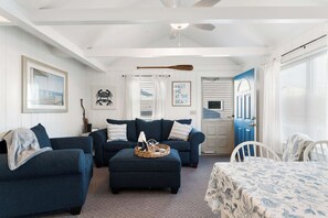 A living room with a blue sofa set, a coffee table with a basket, a ceiling fan, and a door leading to a beach-themed room. - Sand Dollar (Wells)