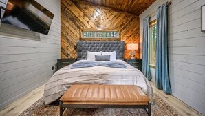 A bedroom with a wooden ceiling, a large bed with a tufted headboard, a bench at the foot of the bed, a TV mounted on the wall, and a window with curtains. - All the Vibes + 2 Story + Shuffle Board + Split Floor Plan + Ride to ATV Trails (Broken Bow)