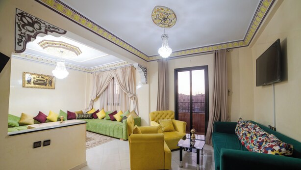 Un salon avec un canapé vert, un fauteuil jaune et un divan vert agrémenté de coussins colorés. Un téléviseur est fixé au mur et un lustre est suspendu au plafond.