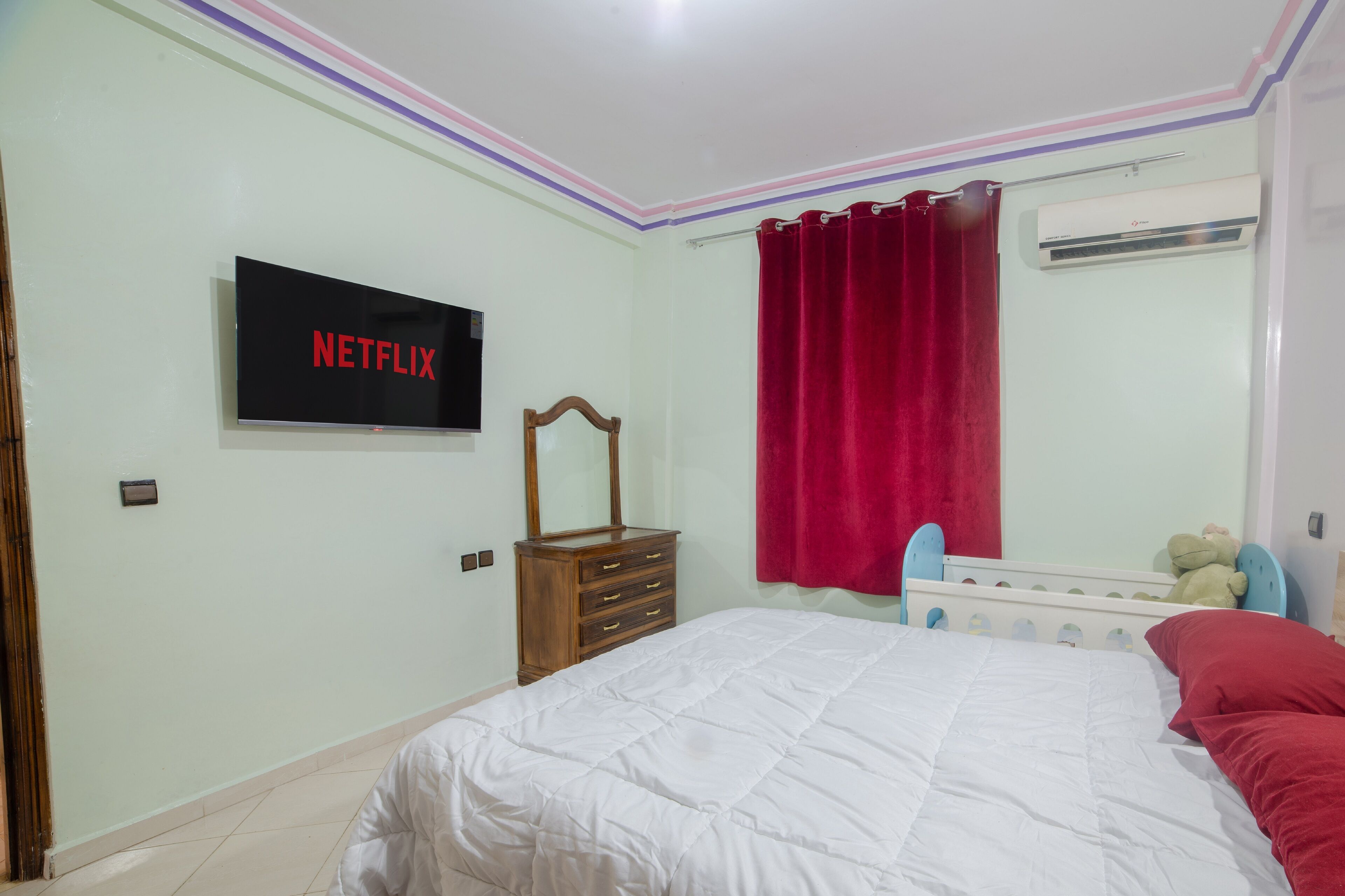 A bedroom with a bed, a wooden dresser, a TV mounted on the wall, a red curtain, and a window with an air conditioning unit.