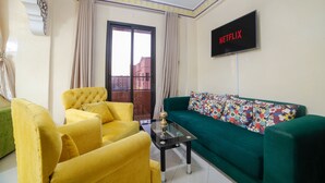 Un salon avec un canapé vert, des fauteuils jaunes, une table basse en verre et un téléviseur à écran plat fixé au mur.