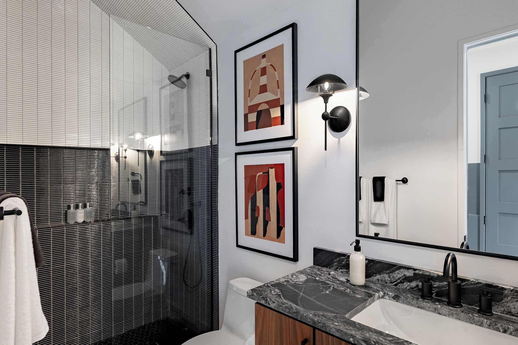 A modern bathroom with a glass shower enclosure, a large mirror, and framed artwork on the wall.
