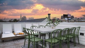 Eine Dachterrasse mit einem weißen Tisch und grünen Stühlen mit Blick auf eine Skyline bei Sonnenuntergang.