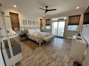Quarto com cama, um banquinho, mesa de cabeceira e vista para a praia através de uma porta deslizante.