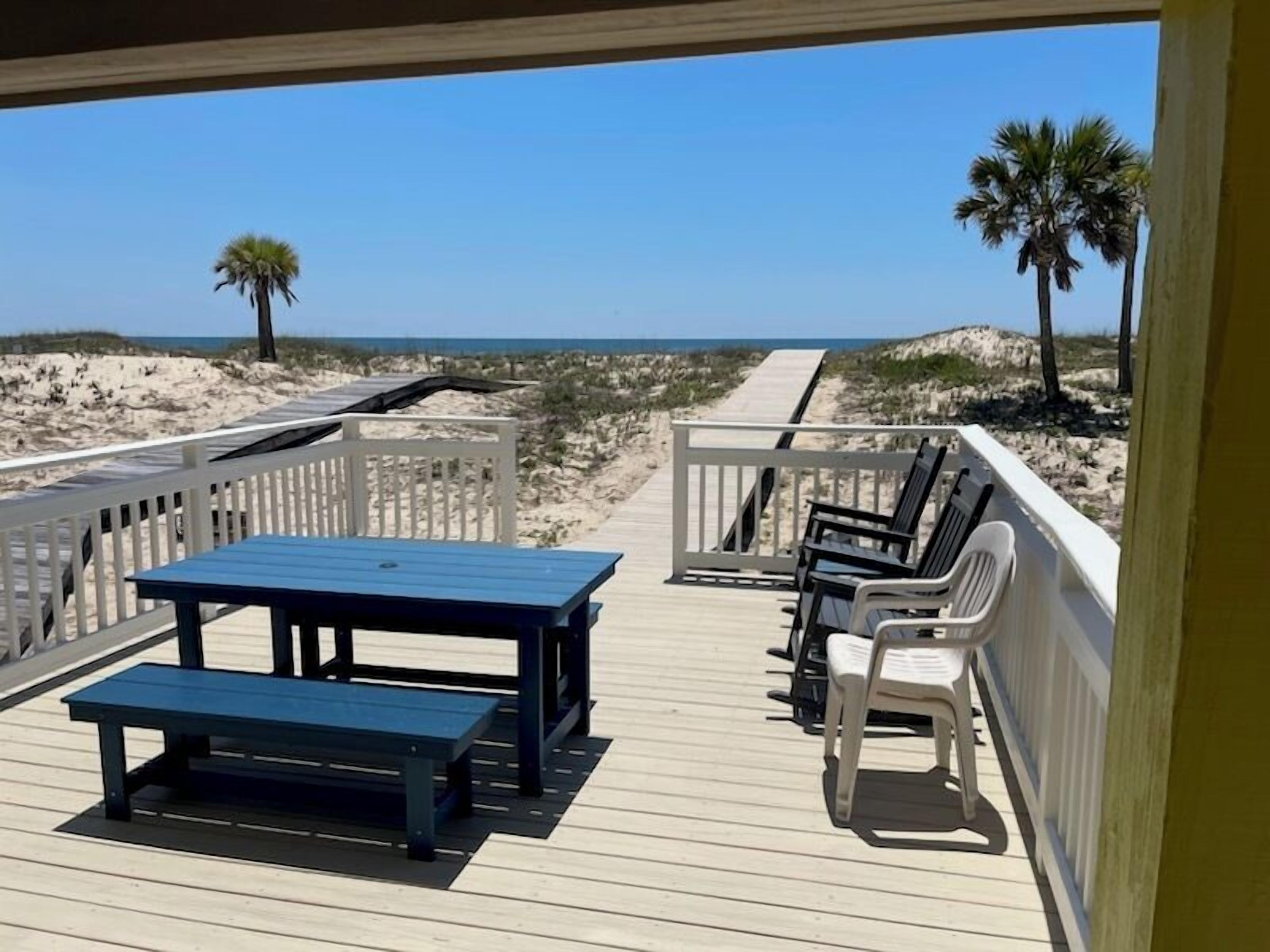 Une terrasse en bois avec une table et des bancs, donnant sur une plage bordée de dunes de sable et de palmiers.