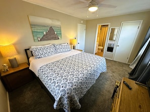 Ein Schlafzimmer mit einem Bett, Nachttischen, einer Lampe, einem Deckenventilator und einer Tür zu einem anderen Raum.