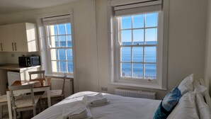 A room with a bed, a desk, a chair, and a view of the sea through large windows. - The Horizon Sea View Suite (Weymouth)