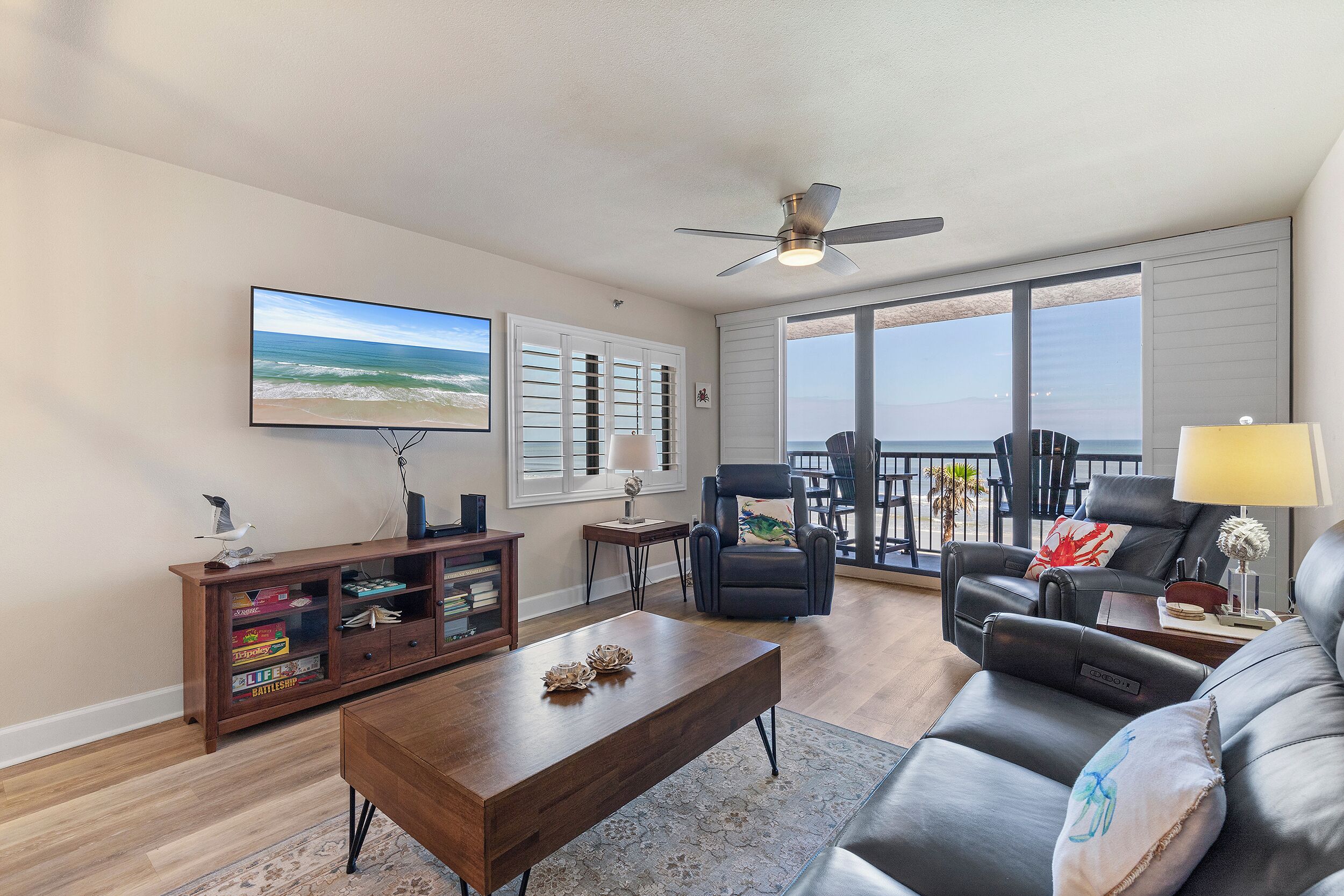 A spacious living room with a wooden coffee table, leather sofa, and a large TV. There is a balcony with a view of the ocean.