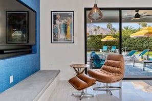 Een moderne kamer met een groot raam, een ingelijst schilderij, een bruine leren fauteuil en een kleine ronde tafel.