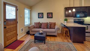A cozy living room with a sofa, ottoman, and coffee table, a kitchen with dark cabinets and a microwave, and a wooden door. - Bluebird Day - Twin Mills Bungalow in Bend, OR, featuring 2 cozy bedrooms. (Bend)