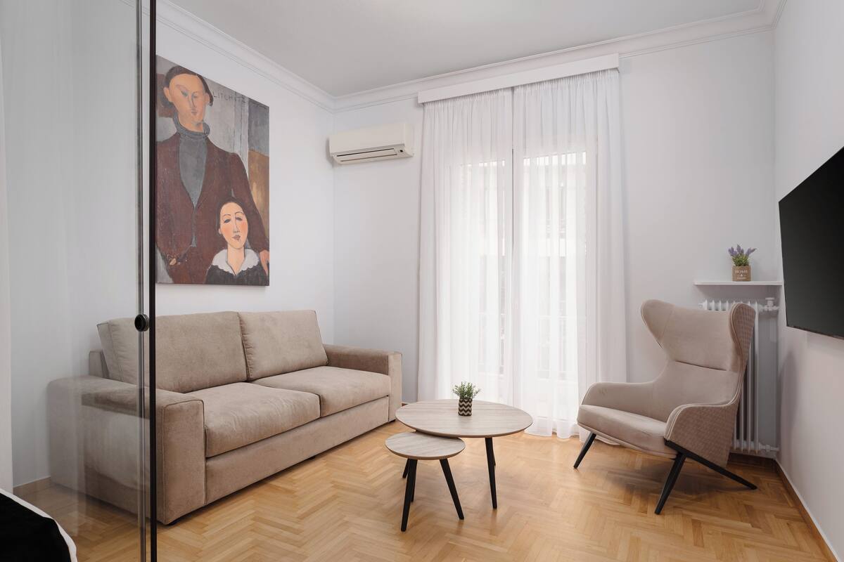 Un salon avec un canapé beige, une table basse ronde, un fauteuil beige, un tableau au mur et une grande fenêtre avec des rideaux.