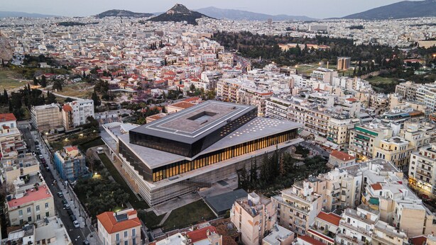 A modern building with a flat roof and large windows, surrounded by a dense urban area with various buildings and a mountain in the background. - Acropolis Classy Apartments (Athens)