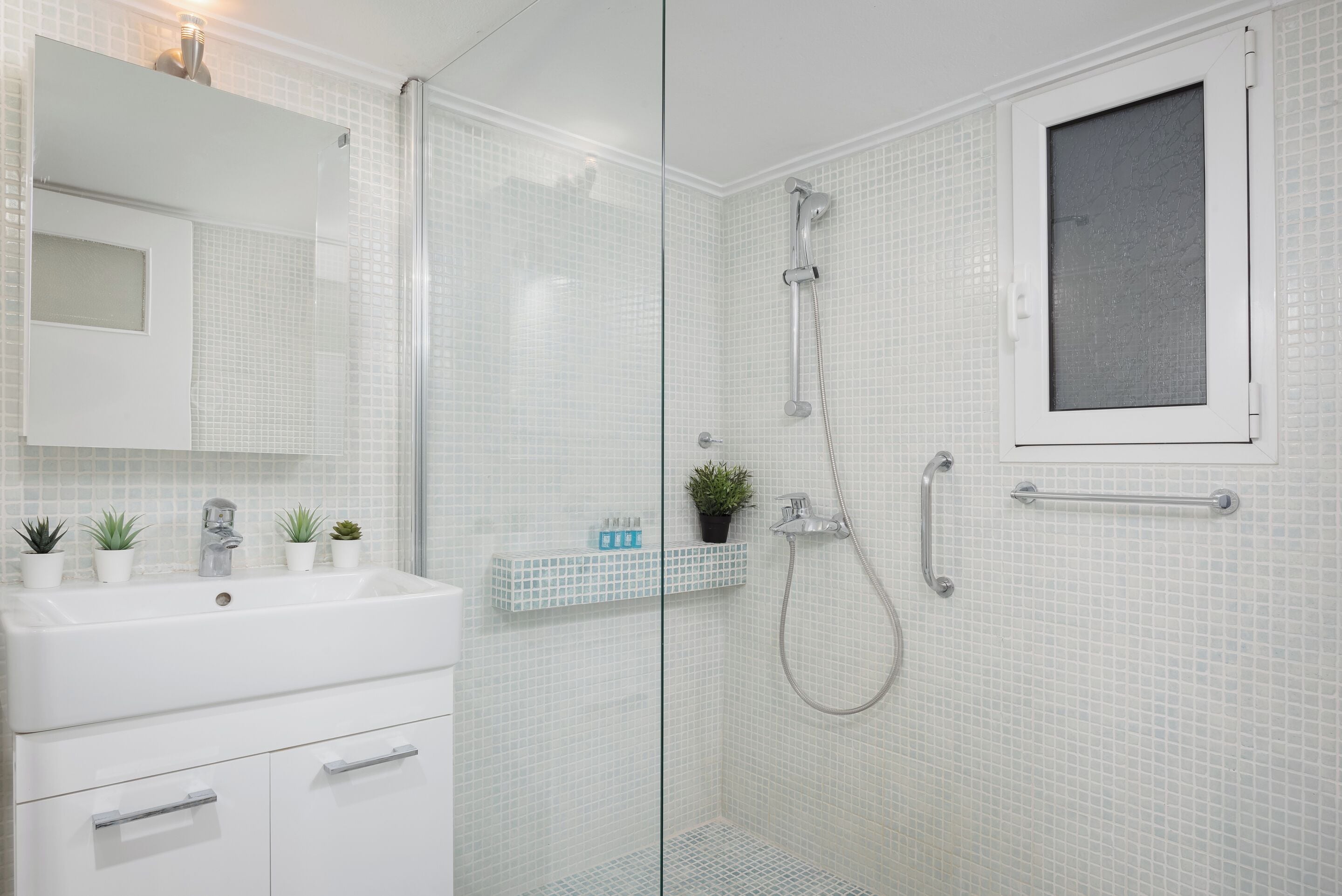 Un baño moderno con cabina de ducha de vidrio, lavamanos blanco y una pequeña ventana.