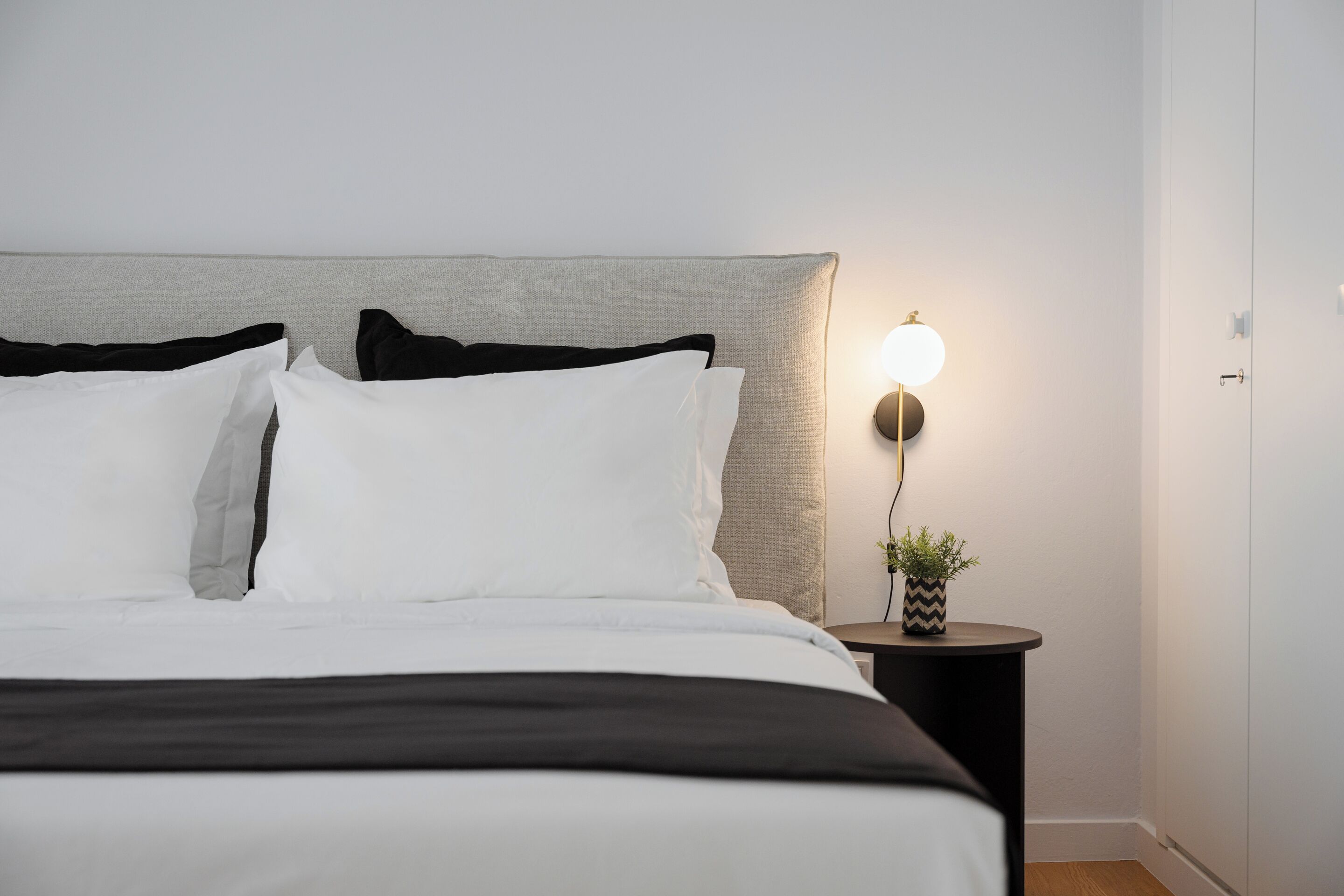 A neatly made bed with white and black bedding, a round bedside table with a lamp, and a small potted plant.