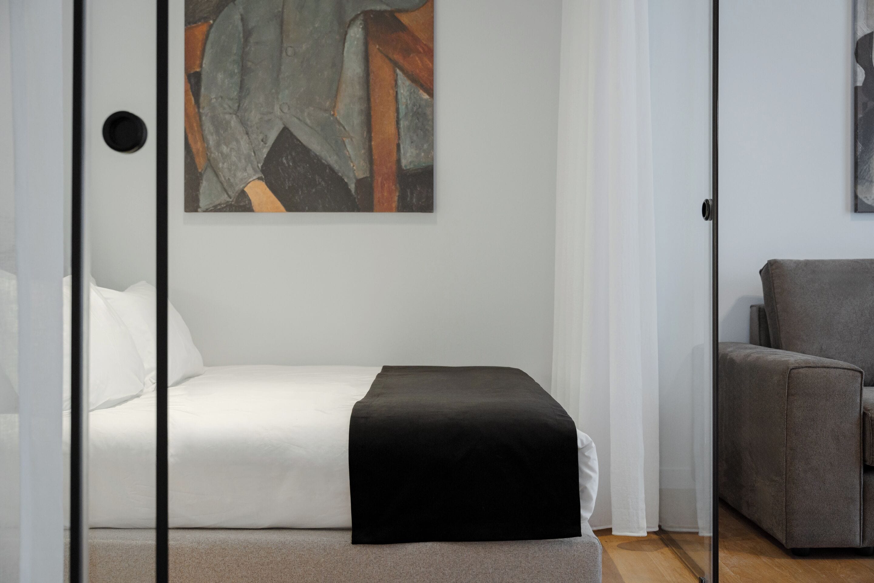 A modern bedroom with a bed, a black throw, a gray sofa, and a geometric abstract painting on the wall.