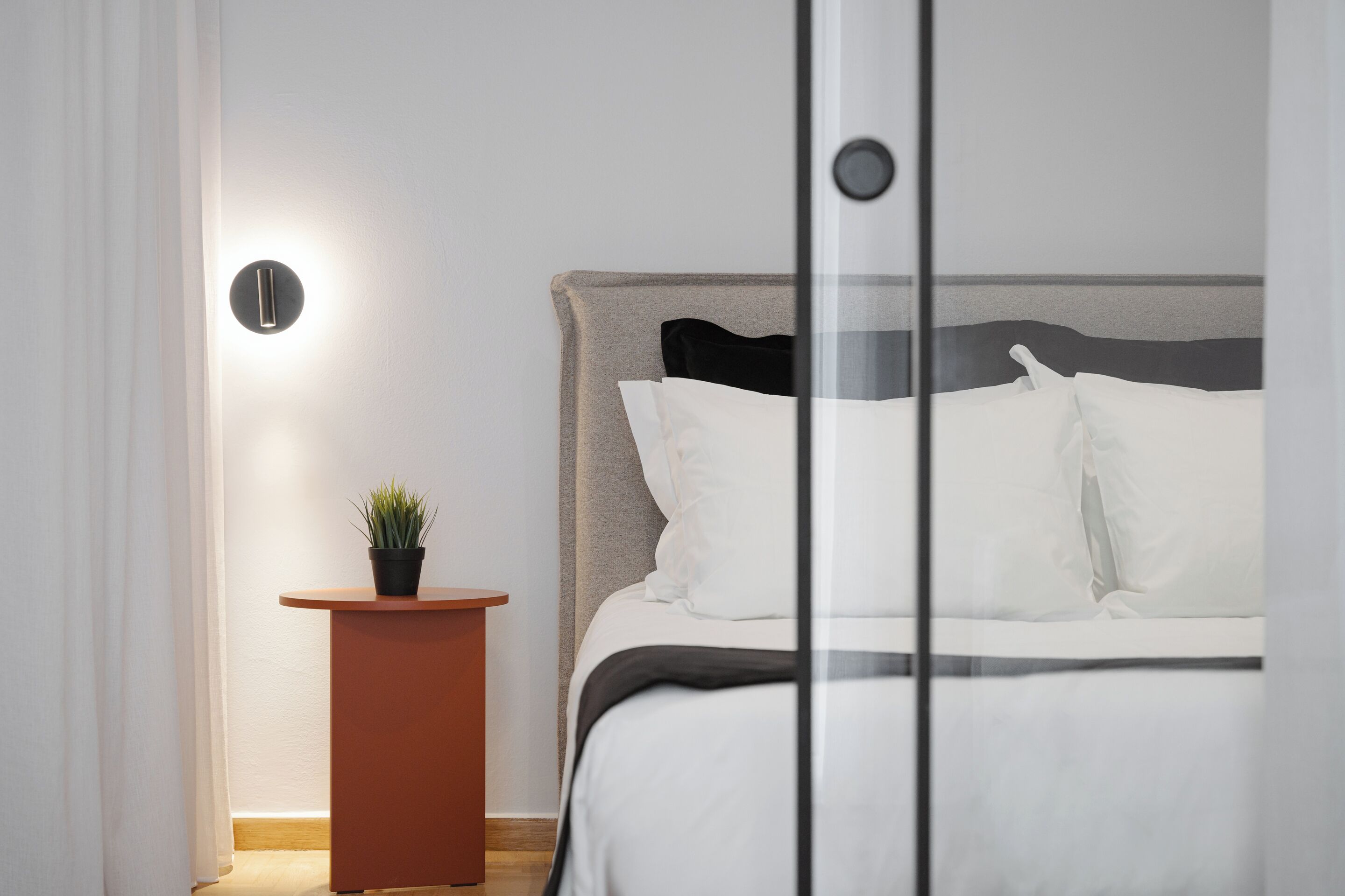 A neatly made bed with white linens, a small round table with a potted plant, and a wall-mounted light fixture.