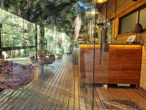 Una terraza de madera con mostrador de recepción, un sofá y una pared de vidrio que ofrece vistas a árboles.