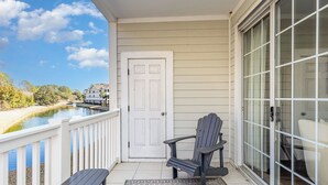 A balcony with a white door, a gray Adirondack chair, a small table, and a view of a body of water with houses in the background. - River Crossing #121- End Unit in Gated Community! (North Myrtle Beach)