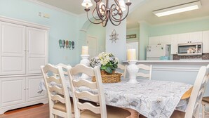 A dining area with a table set for four, surrounded by white chairs with woven seats. A chandelier hangs above, and a basket with flowers is on the table. - River Crossing #121- End Unit in Gated Community! (North Myrtle Beach)