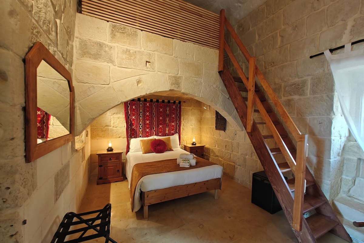 A bedroom with a stone wall, a wooden bed, a nightstand, a mirror, a chair, and a wooden staircase.