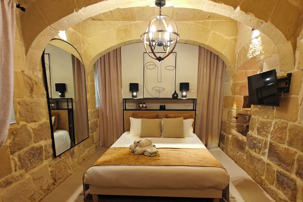 A hotel room with a stone archway, a large bed with a headboard, a chandelier, a flat-screen TV, and a mirror.