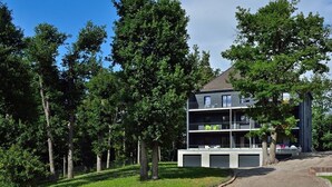 Een modern, meerhoog gebouw met balkons, omringd door bomen en een grindpad.