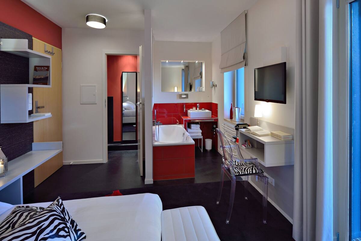 Una habitación de hotel moderna con cama, baño con pared de acento roja, televisor y ventana.