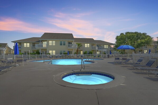 A pool area with two circular pools, lounge chairs, and a building with balconies in the background.