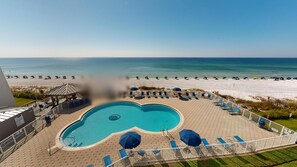 Zona de piscina con sillones y sombrillas, con vista a una playa que también tiene sombrillas y gente.