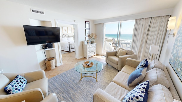 Un salón amplio con una mesa de centro de vidrio, sofás beige con cojines estampados, un televisor de pantalla plana y vistas al océano a través de ventanas grandes.