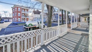 A covered porch with a white lattice railing, a concrete mixer truck parked outside, and a row of colorful townhouses in the background.