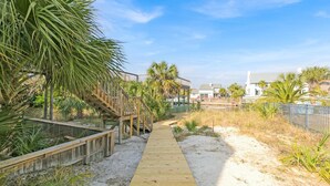 A wooden boardwalk leading to a wooden deck with a railing, surrounded by sand and vegetation, with buildings in the background.