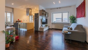 Eine moderne Wohnung mit Küche, Essbereich und Wohnzimmer. Es gibt Holzböden, ein graues Sofa und ein großes Fenster mit Blick auf die Stadt.