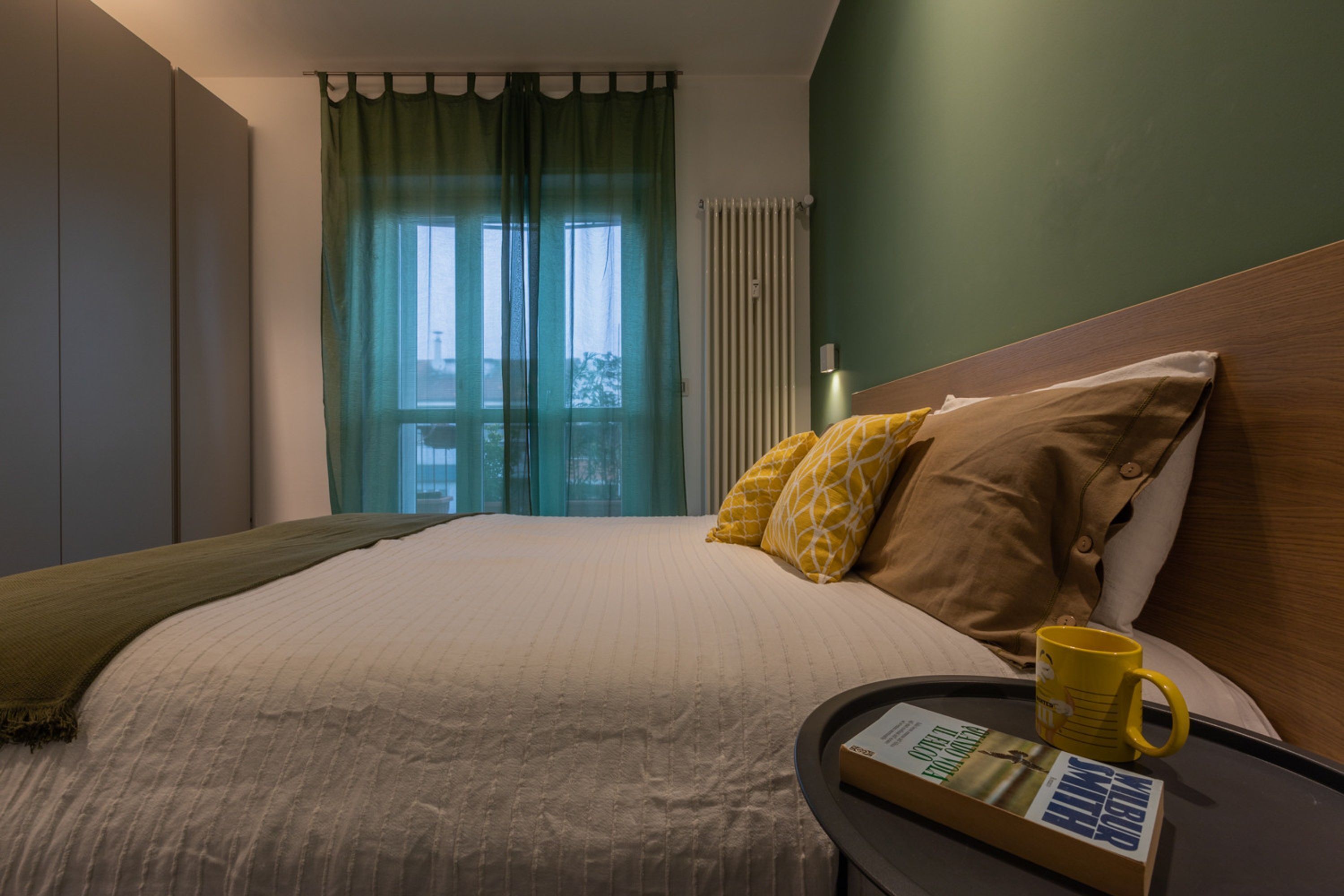 A neatly made bed with a white and beige duvet, two pillows with yellow and brown patterns, a yellow mug, and a book on a side table.