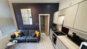 Une cuisine moderne avec des meubles gris, une cuisinière noire et un four à micro-ondes. Le salon est doté d’un canapé gris agrémenté de coussins jaunes.
