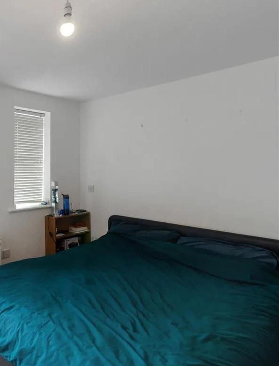 A bedroom with a bed, a window with blinds, and a small wooden shelf with items.