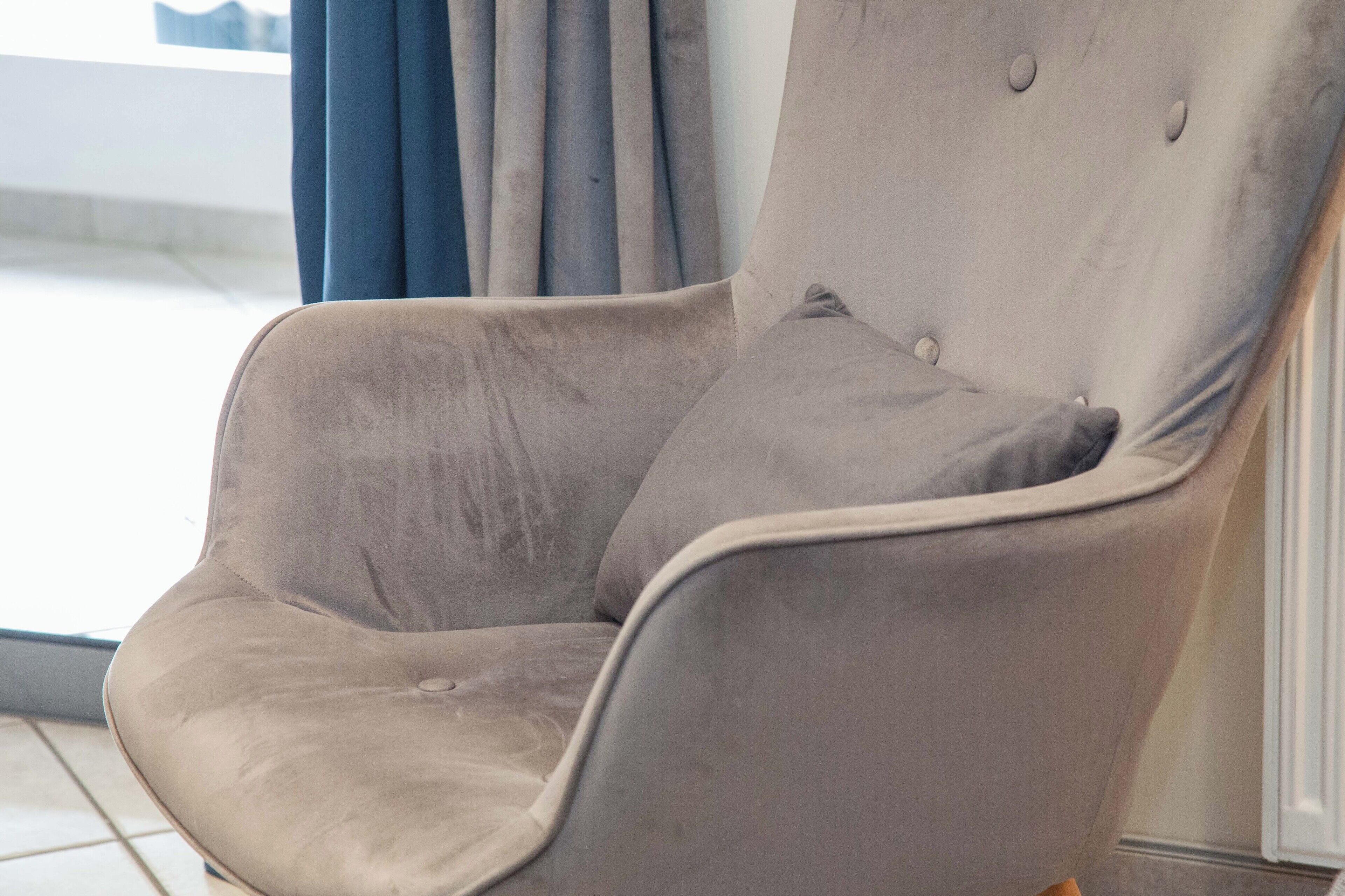 A grey upholstered armchair with a cushioned backrest and armrests, positioned near a window with curtains.