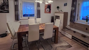 A modern dining area with a wooden table and white chairs, a ceiling light fixture, and a window with curtains. - Vacation home "Green Emotion" (Suhl)