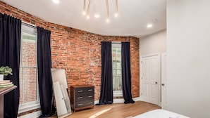 A bedroom with a brick wall, a wooden dresser, a bed with white linens, and a mirror leaning against the wall. - Access OSU & Crew Stadium | Pet Friendly | Parking (Columbus)